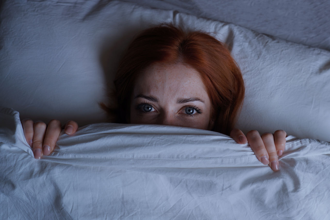 Sleepless woman lying in bed hiding under duvet at night