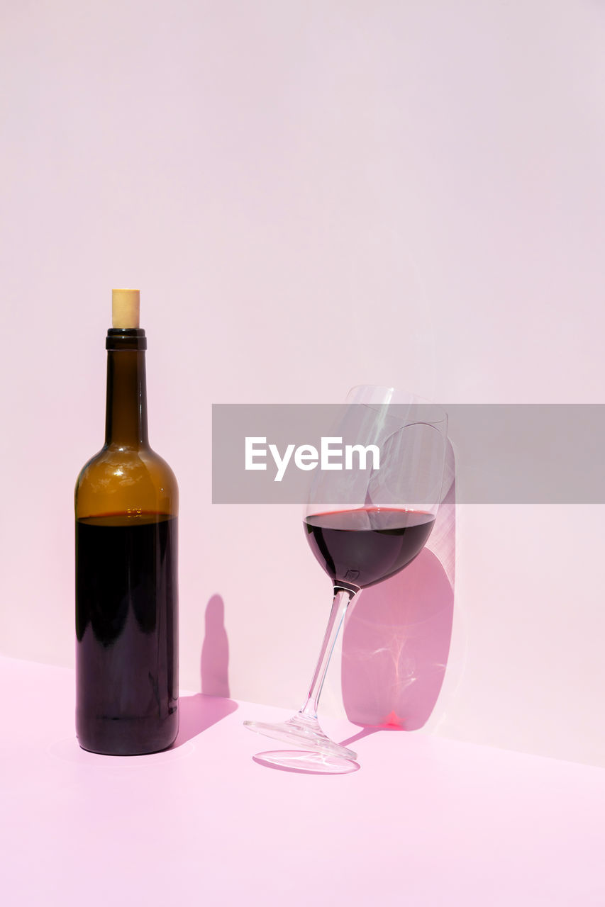 A bottle of red wine and a filled glass next to it in the sun on a pink background. place for text