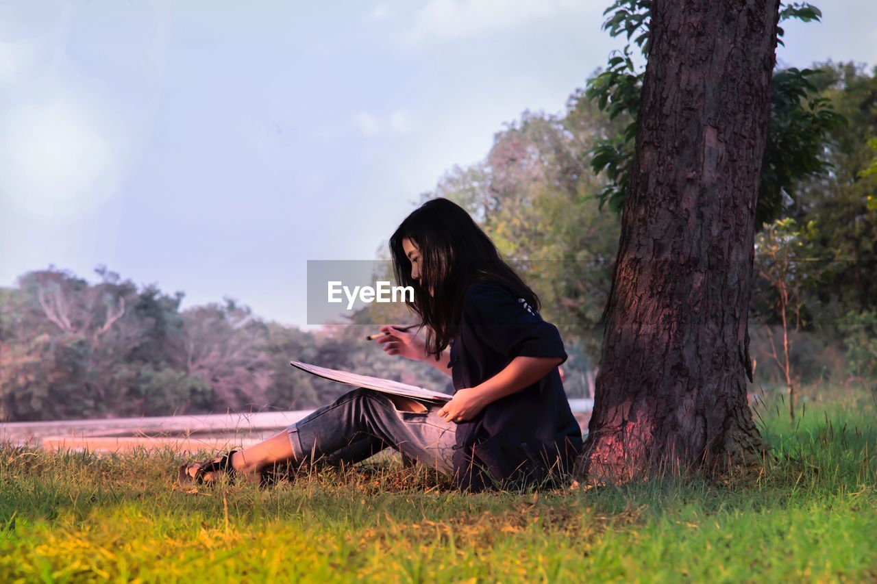 Side view of woman painting while sitting by tree trunk on field