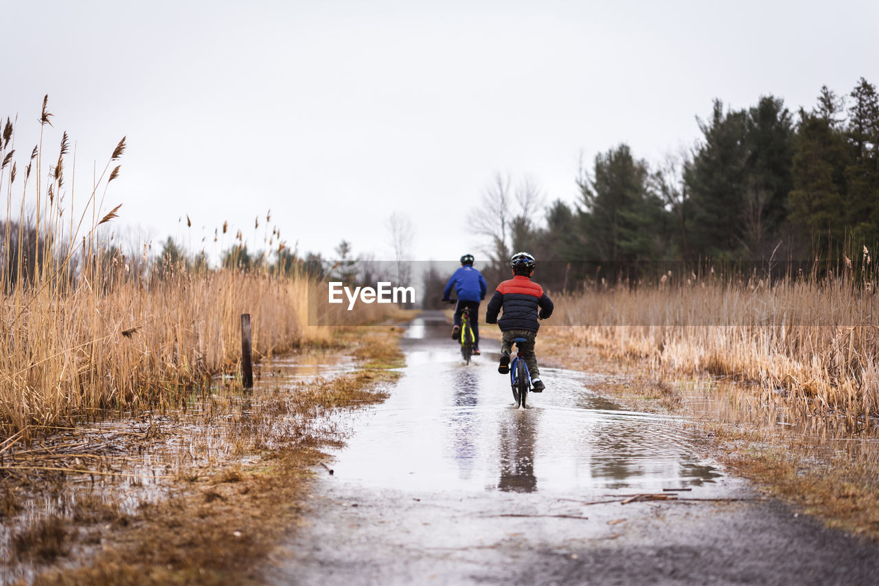 Two boys riding their bikes through a large puddle on a flooded trail.
