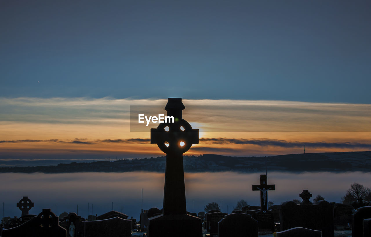 Cross and tombstones in graveyard against sky during sunset