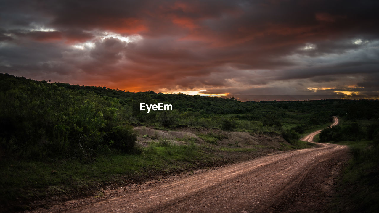 Road by field against cloudy sky during sunset