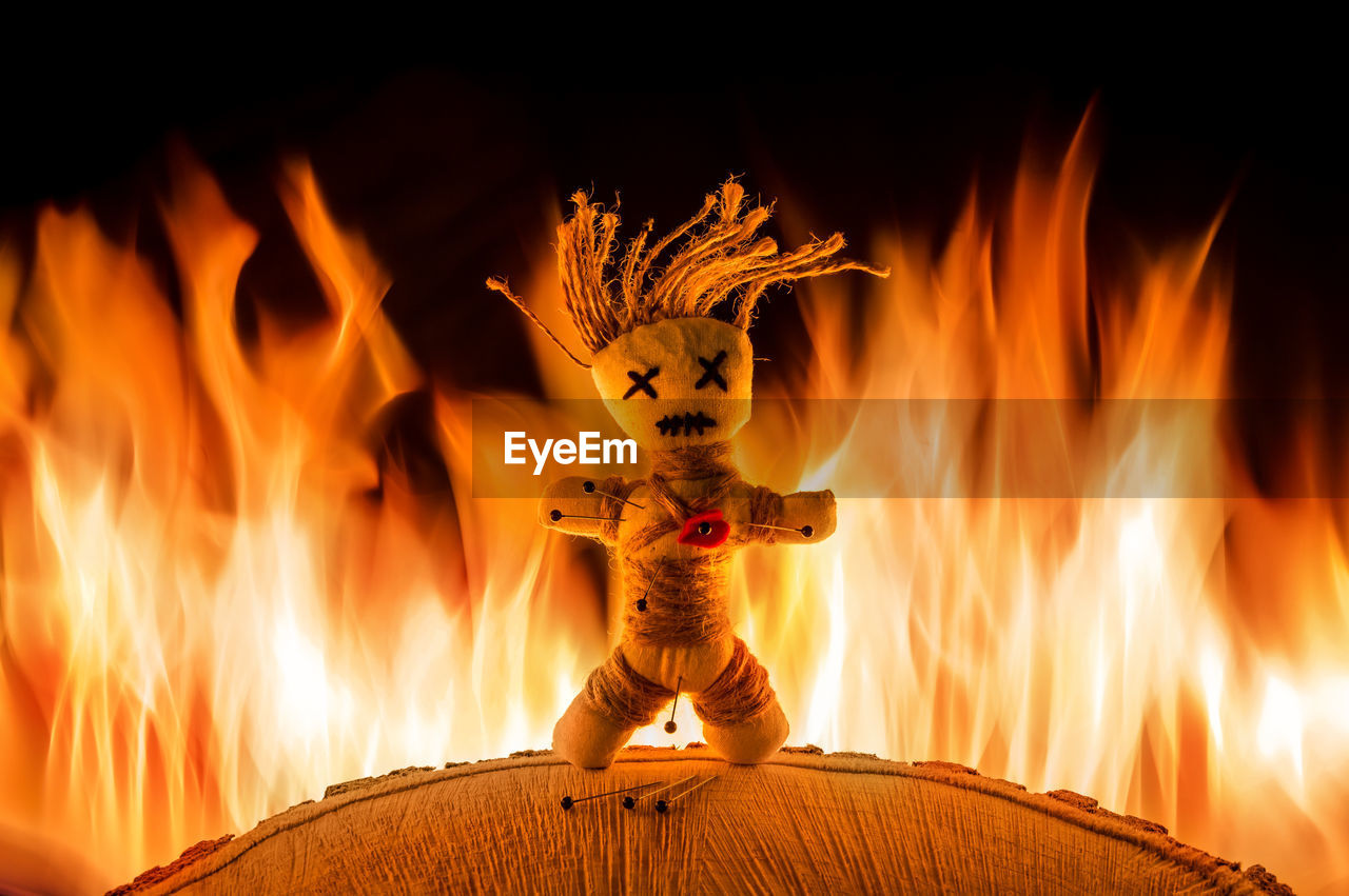 A needle punched voodoo doll stands in front of a bright fire