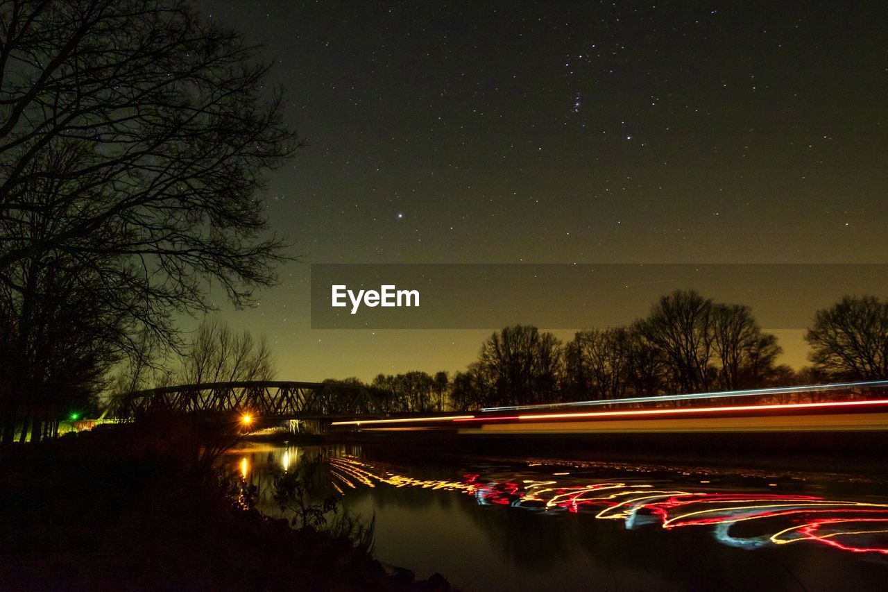 LIGHT TRAILS ON RIVER BY TREES AGAINST SKY AT NIGHT