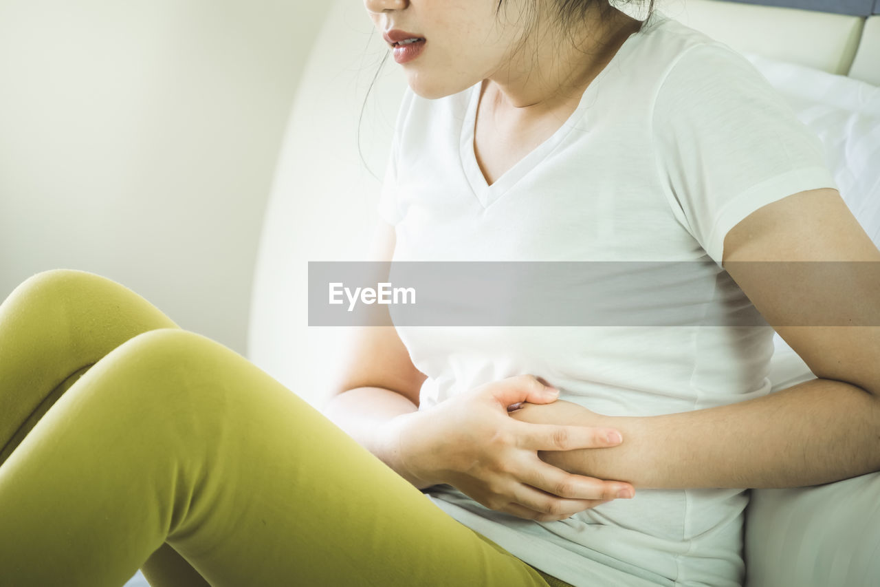 Midsection of woman with stomachache sitting on bed at home