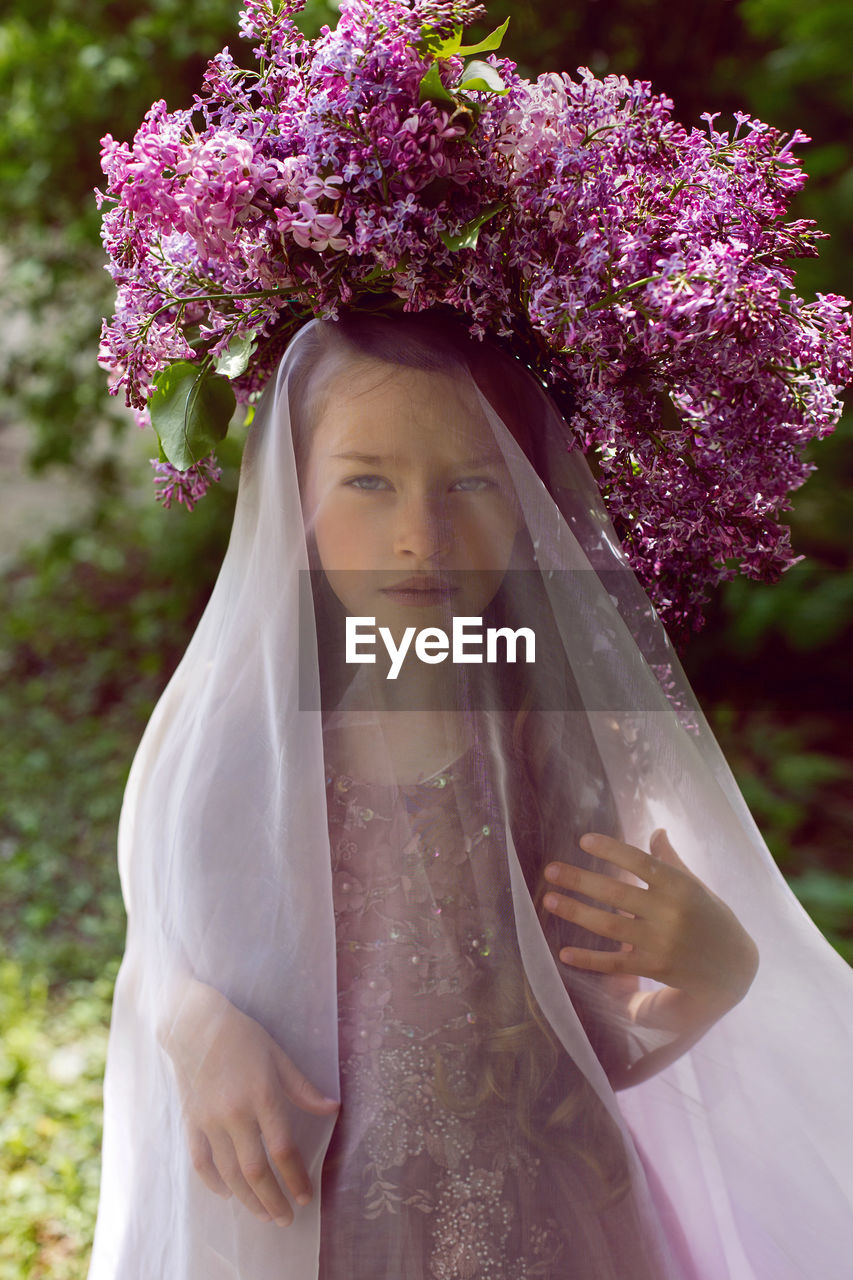 Caucasian girl child seven years old in a purple dress stands in nature with a wreath of lilacs
