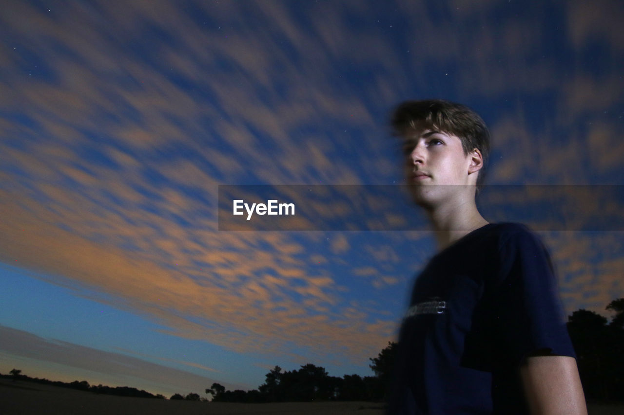 Low angle view of thoughtful teenage boy standing against sky during sunset