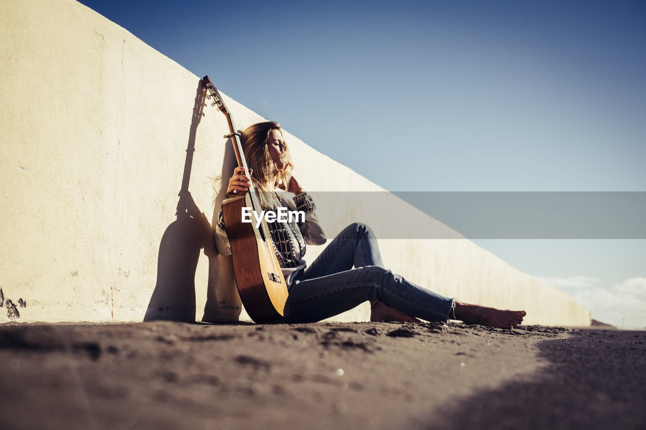 Full length of young woman with guitar leaning on retaining wall against clear sky