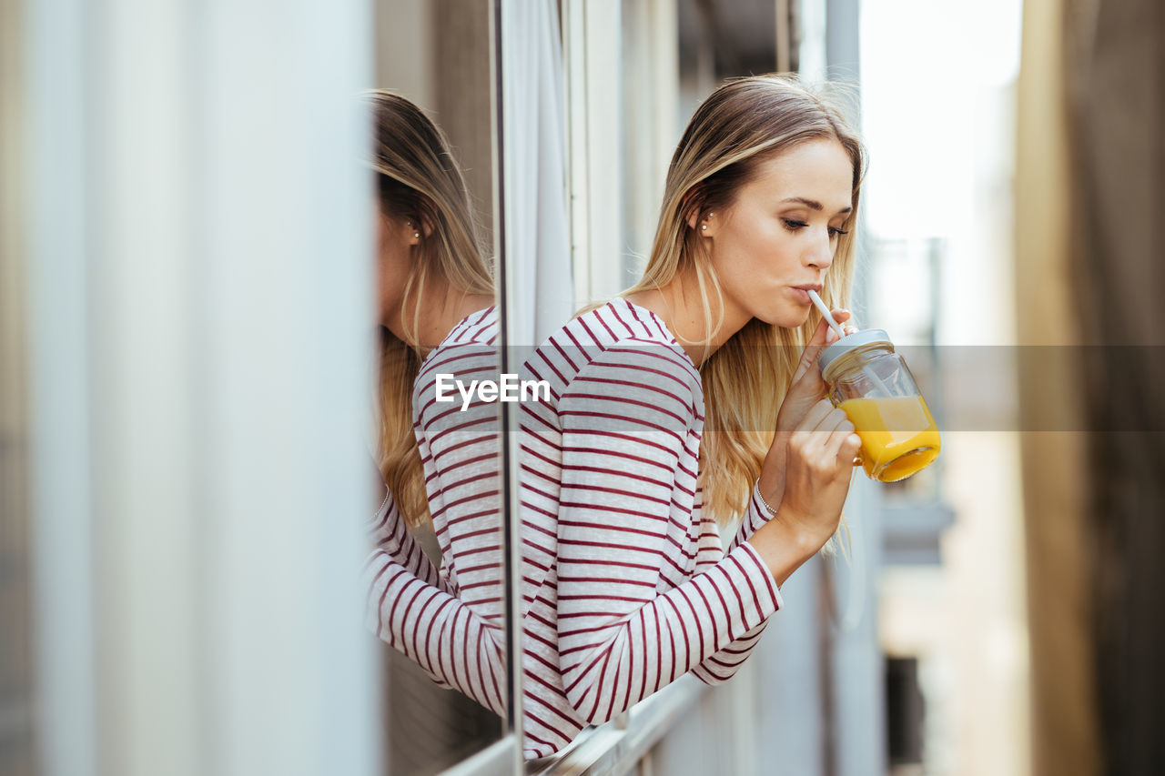 Young woman drinking juice while leaning out of window