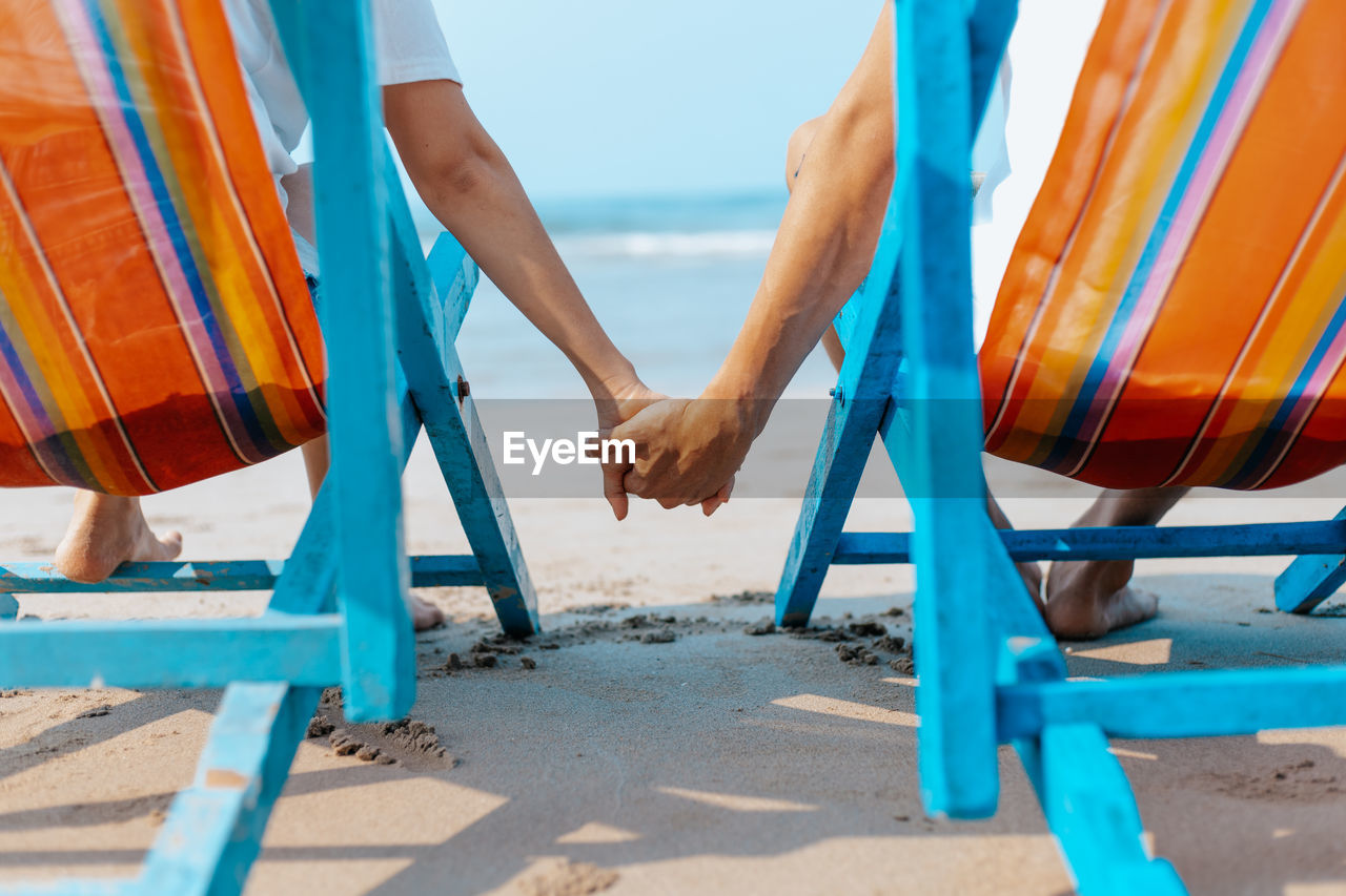 Couple sitting at beach on chair while holding hands at beach