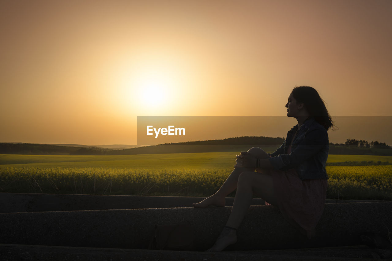 Side view of thoughtful woman looking at view while sitting on field against sky during sunset