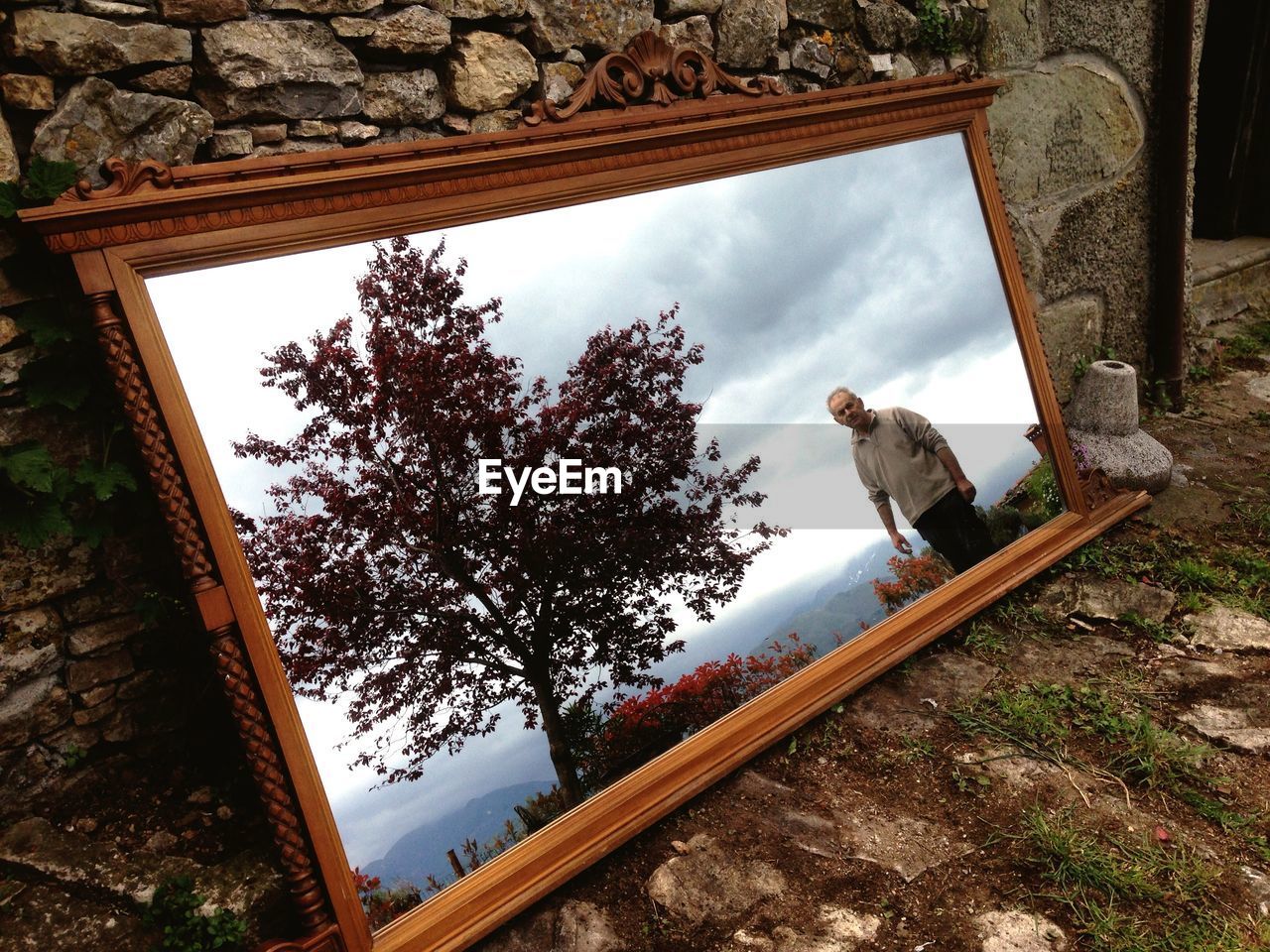 Reflection of mature man and tree on mirror leaning on wall at field