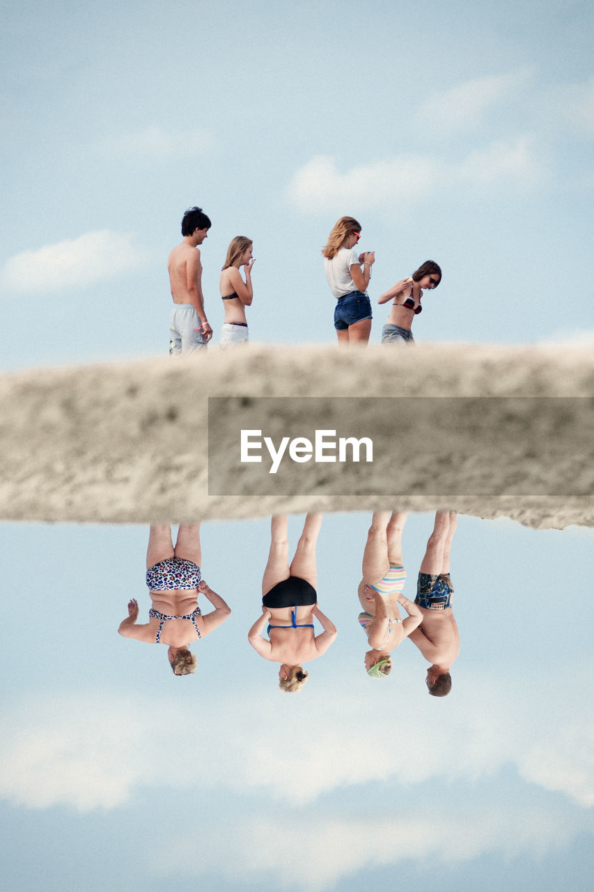 People with reflection in sea standing against sky