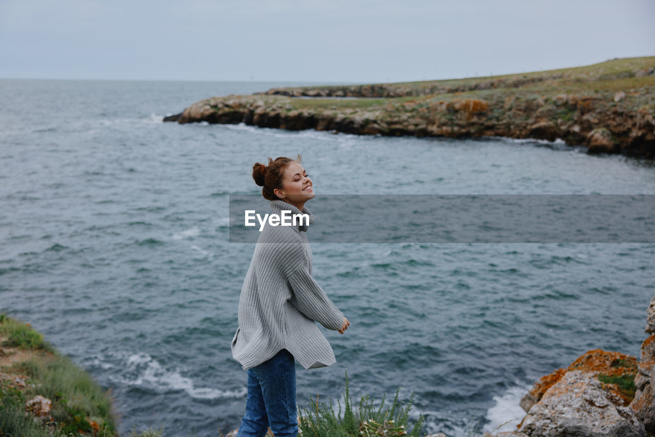 rear view of young woman standing on rock by sea against sky
