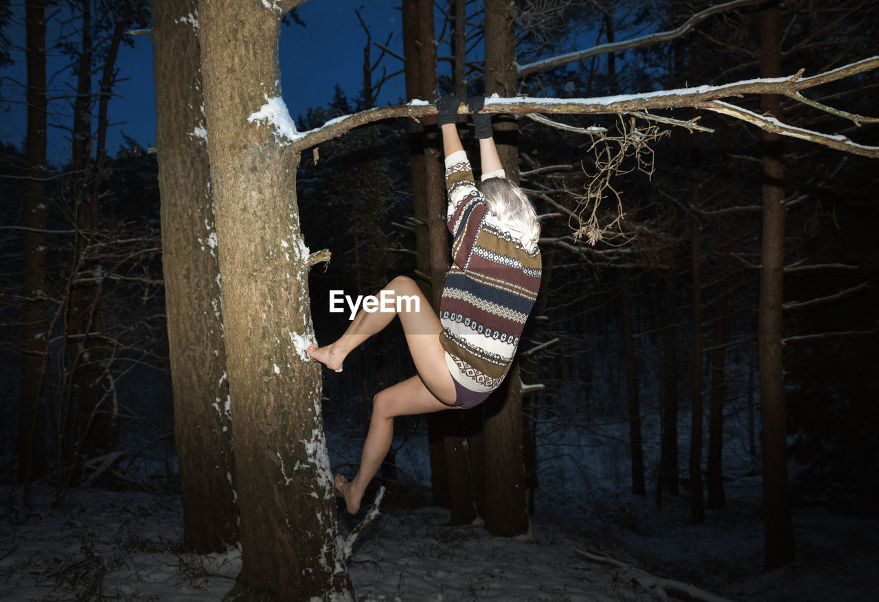 Side view of young woman climbing on tree in forest during winter at night 