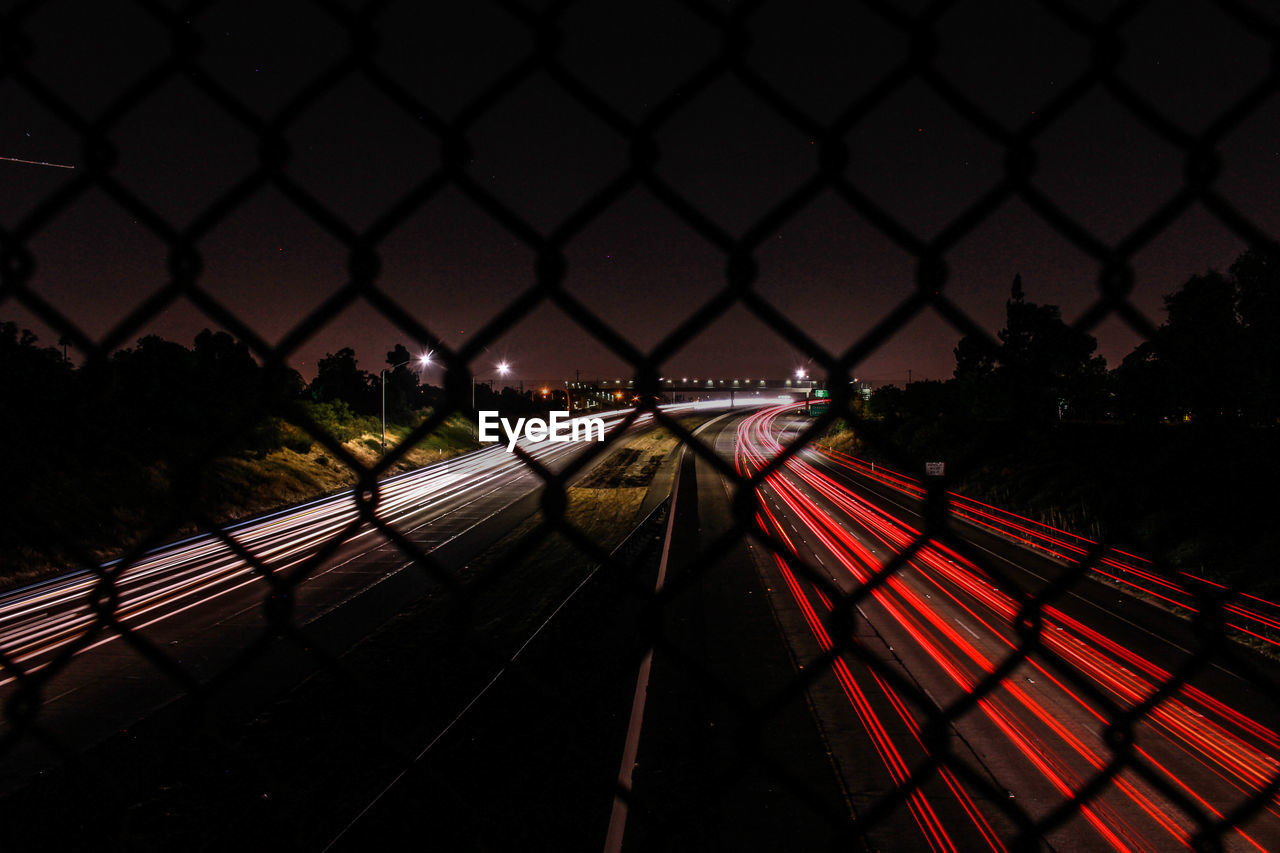 Light trail on highway against sky seen from chainlink fence