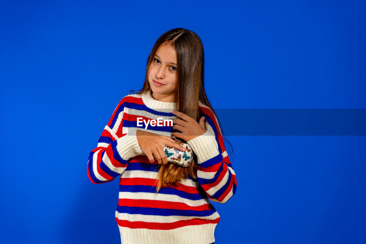blue, one person, colored background, striped, studio shot, women, blue background, copy space, portrait, emotion, child, young adult, person, adult, clothing, waist up, holding, happiness, indoors, long hair, smiling, hairstyle, casual clothing, childhood, standing, photo shoot, front view, looking, looking at camera, brown hair, female, lifestyles, three quarter length, teenager
