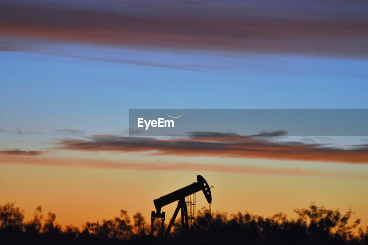 Silhouette oil pump amidst trees against orange sky during sunset