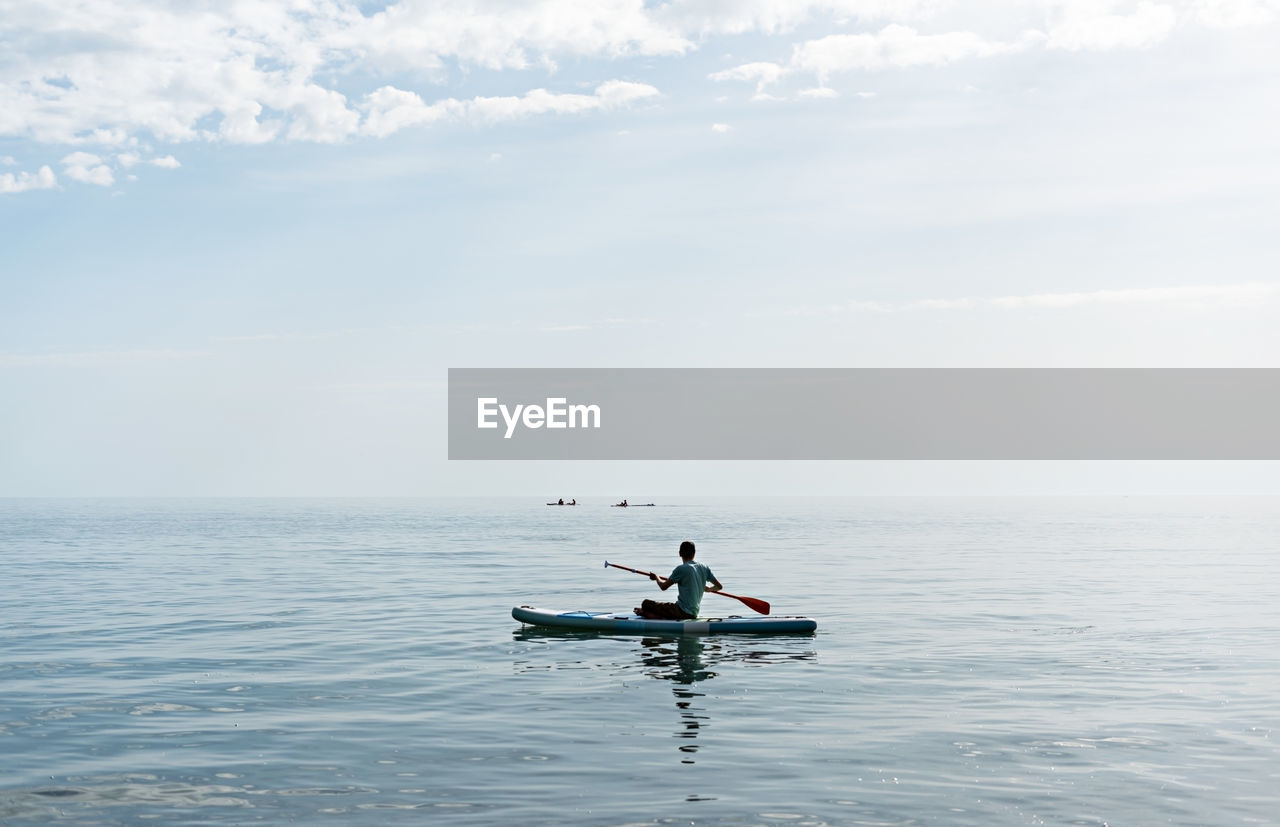 Young man from behind sitting on stand up paddle board on sea on sunny summer day, active lifestyle