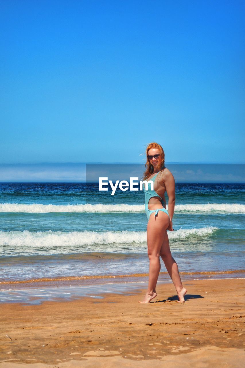 Full length of young woman tiptoeing on shore at beach against clear blue sky