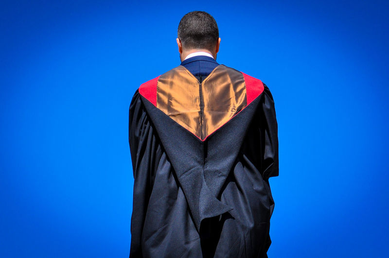 Rear view of man in graduation gown standing against clear blue sky