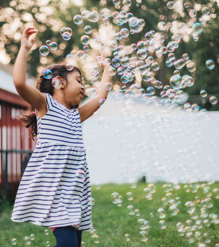 Diverse mixed race pre school girl outdoors during summer having fun in backyard with bubbles 