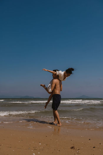 Shirtless man carrying woman while standing at beach against clear sky