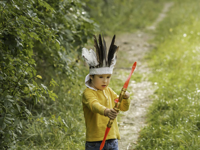 Boy is playing american indian. kid has headdress made of feathers,bow with arrows. costume play. 
