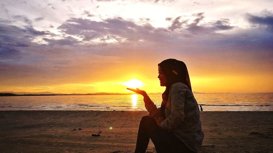 Silhouette woman sitting on beach against orange sky
