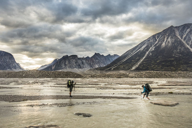 Two backpackers navigate crossing a braided river in a mountain pass.
