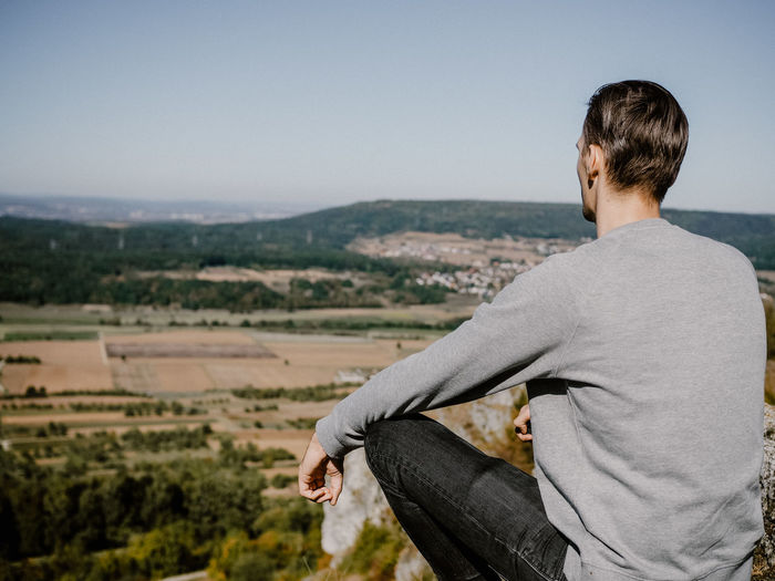 Rear view of man sitting on cliff against sky