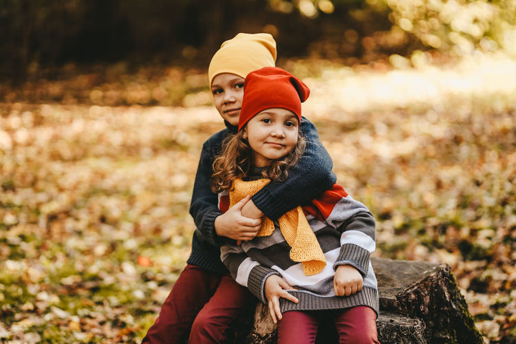 A little girl and a boy children brother and sister hug and sit on a tree stump in the autumn forest