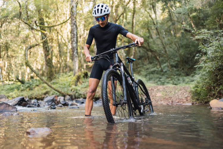 Male bicyclist walking with bike in river with foam during trip in woods