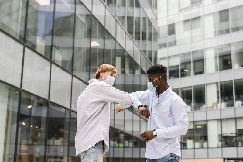 Friends in surgical masks doing elbow bump by modern building
