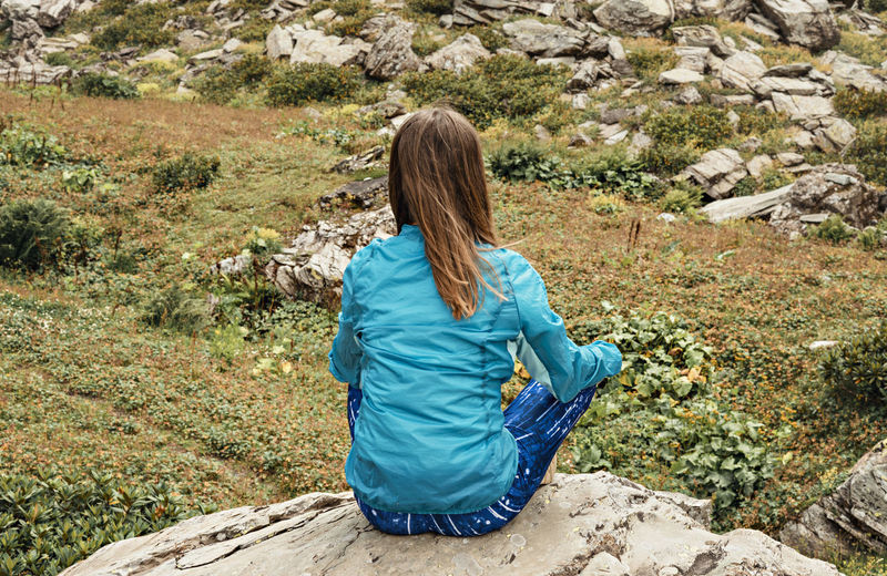 Rear view of meditating young woman in blue sitting on stone in green mountain valley,  mindfulness
