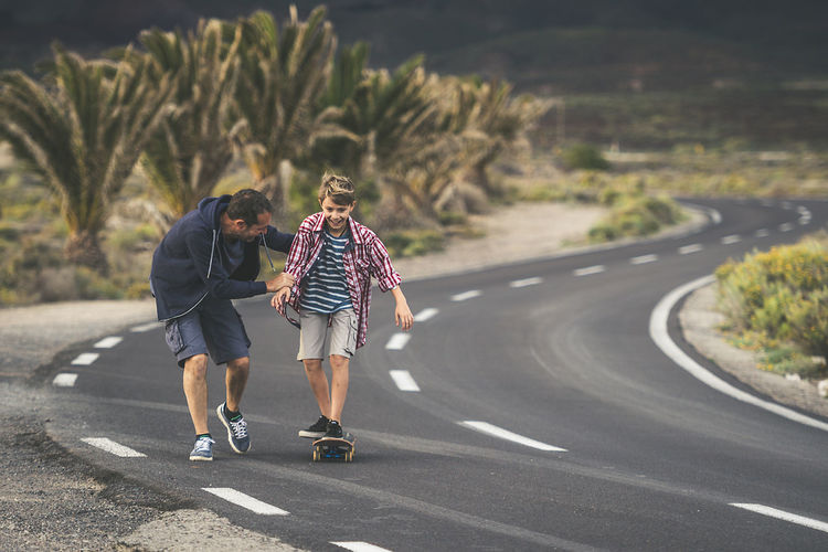 Father assisting son in skateboarding on road against plants
