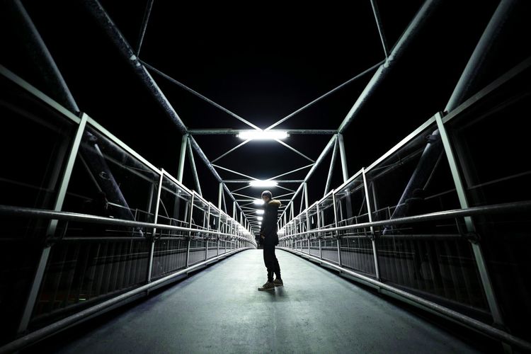 Young man standing on illuminated footbridge against sky at night