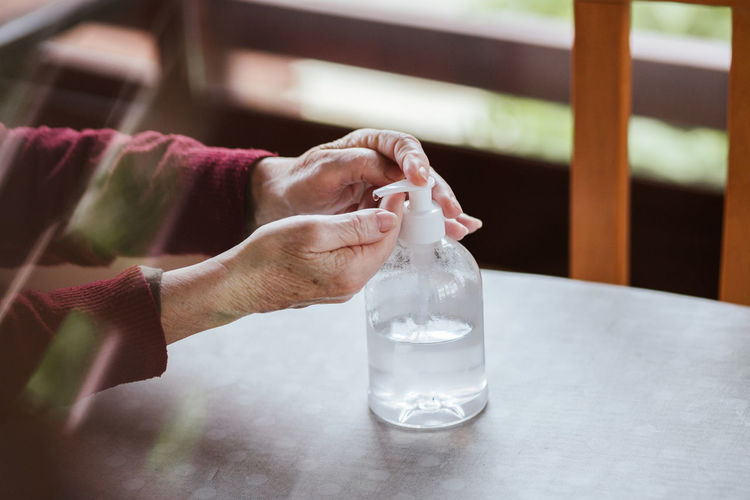 Unrecognizable person with wrinkled hands taking sanitizer from bottle with dispenser while disinfecting hands at home person