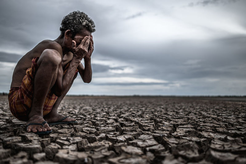 Man crying while crouching on arid land against sky