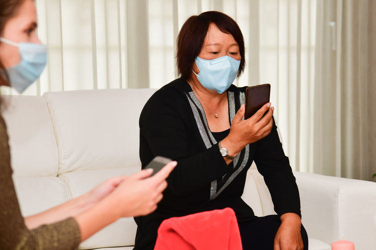 Woman wearing mask using smart phone while sitting on sofa