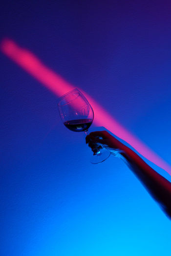 Closeup of woman hand holding glass of wine over fashion blue wall, neon blue stripe on wall.