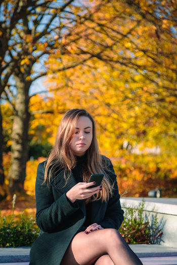 A woman with long hair sits on a bench in an autumn park and looks into a mobile phone. 