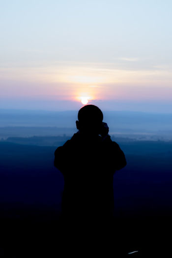 Silhouette man photographing while standing against sky during sunset