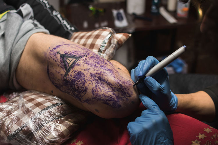 From above crop unrecognizable tattooist in gloves drawing picture with pen on male leg in salon