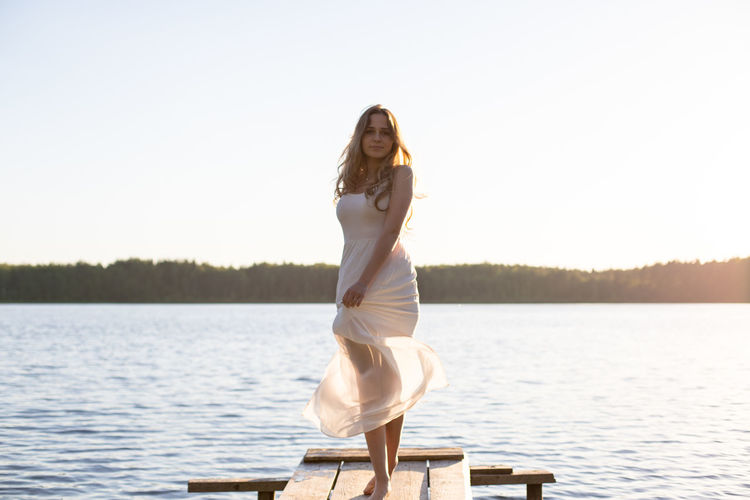 Full length of young woman standing by lake against clear sky