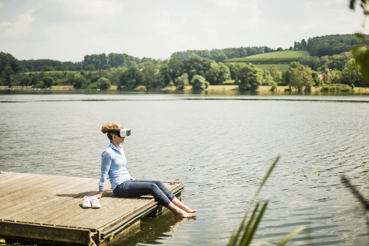 Woman with vr glasses sitting on jetty at a lake with feet in water