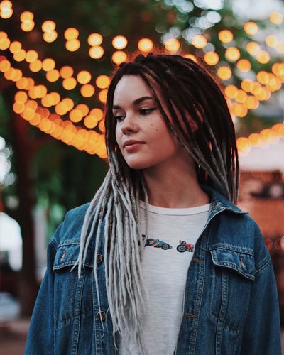 Beautiful young woman with dreadlocks looking away against illuminated lights