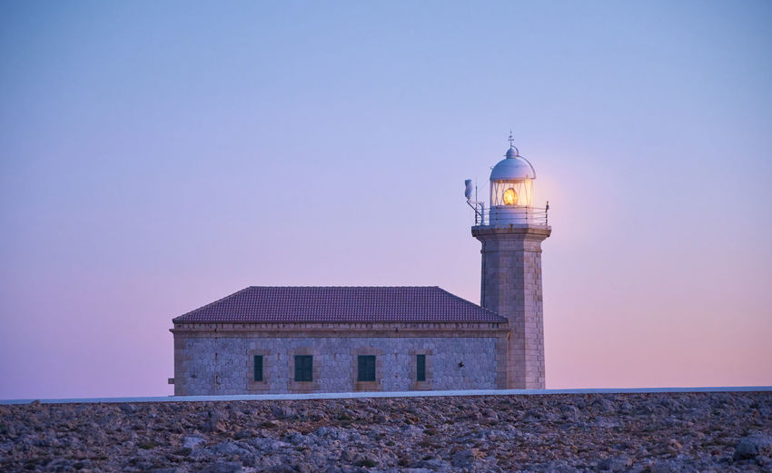 Punta nati lighthouse in menorca turns on at dusk against clear blue and pink sky