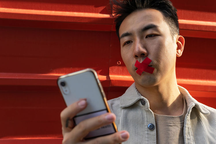Crop unemotional asian male standing near red metal fence with red tape on lips and browsing mobile phone