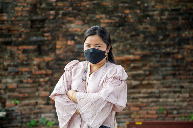 Asian woman in a pink dress cross her arms wearing a face mask with an old brick wall background.
